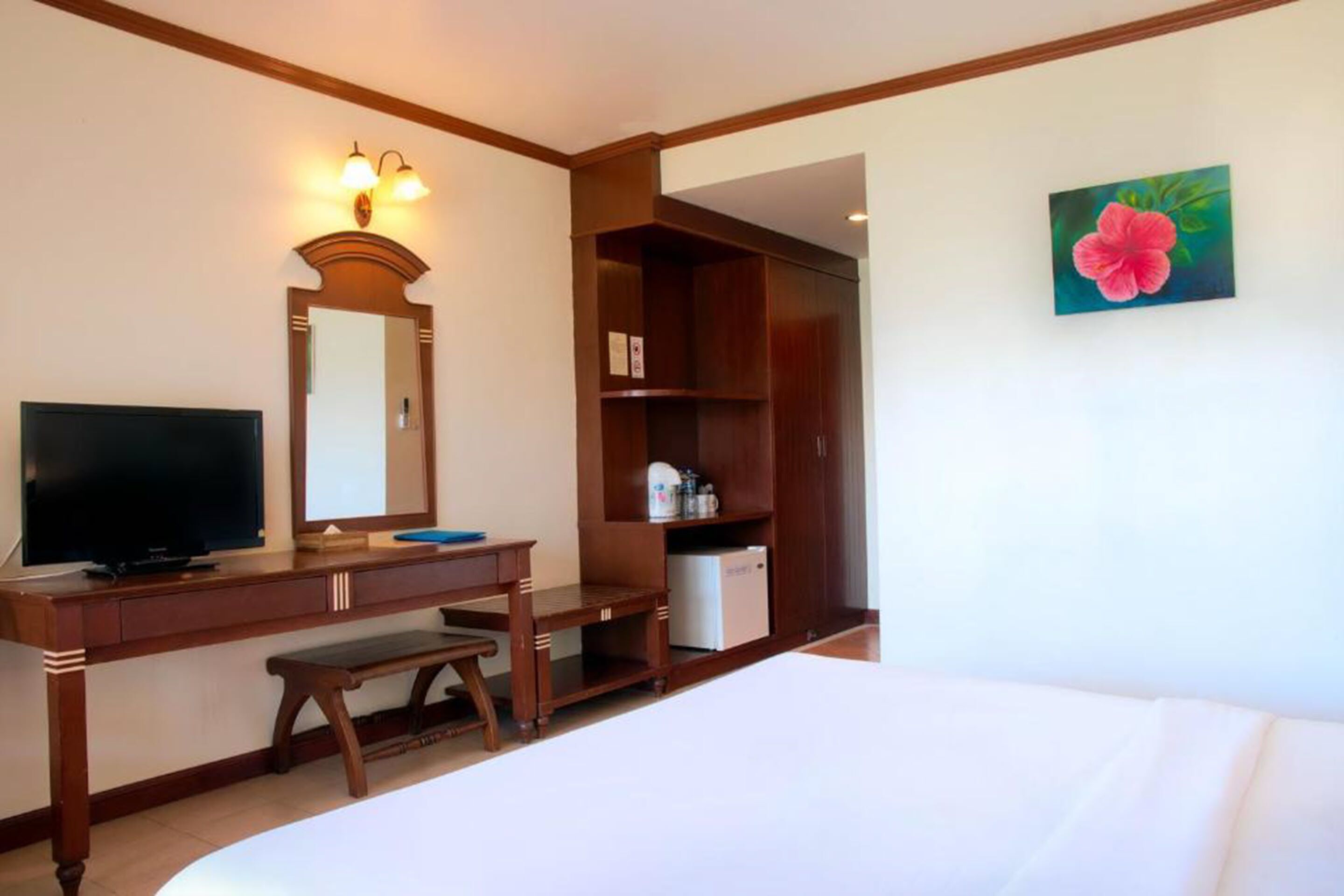 A hotel room with a bed, a desk with a television, a mirror, a shelf with toiletries, and a painting of a flower on the wall.