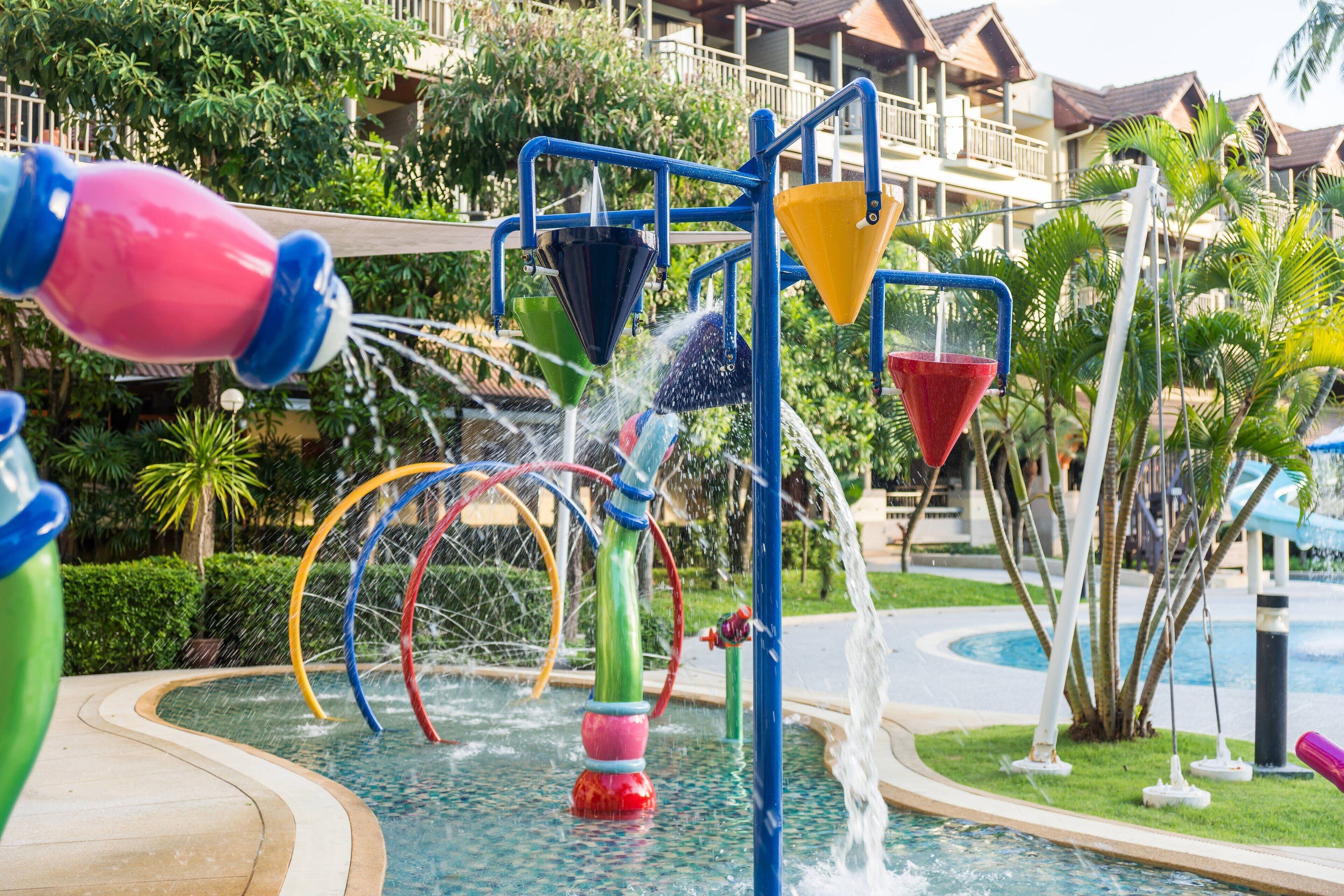 A water play area with colorful slides and fountains, surrounded by lush greenery and a pool.