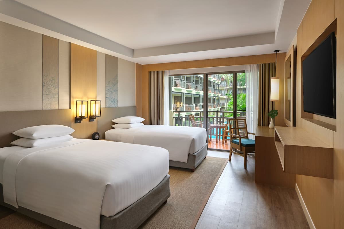A hotel room with two beds, a TV, a desk, and a balcony with a view of the hotel and greenery.