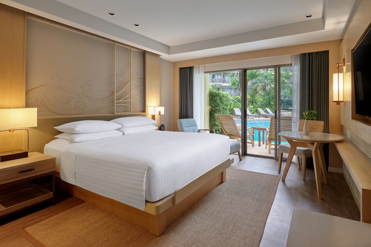 A modern hotel room with a large bed, a desk, a chair, a TV, and a view of a pool and greenery outside.