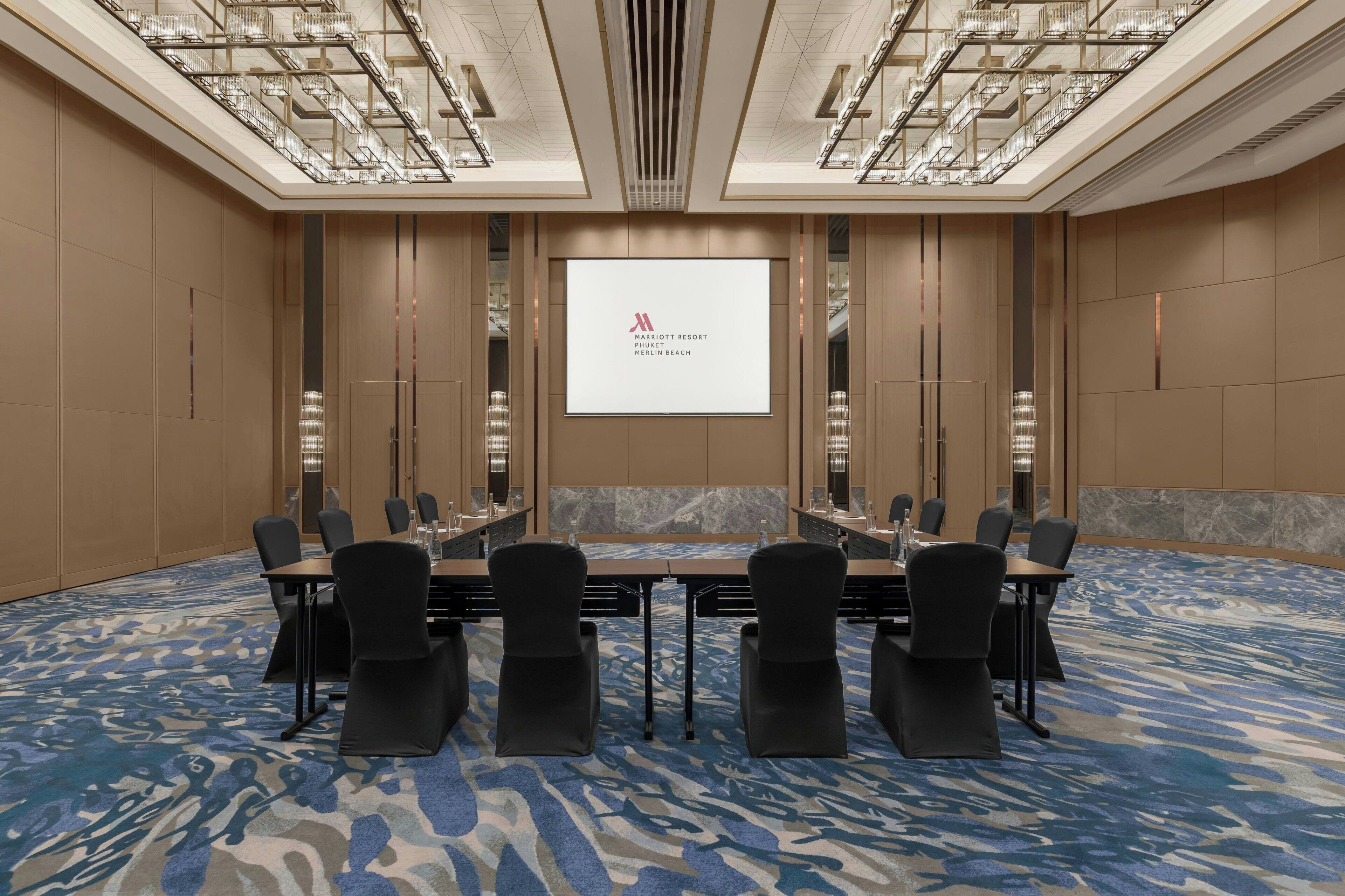 Ein Konferenzraum mit einer großen Leinwand, auf der das Marriott-Logo zu sehen ist, einem langen Tisch mit Stühlen und einem gemusterten Teppich.