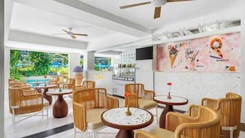A modern indoor seating area with wicker chairs and round tables, a ceiling fan, and a wall with ice cream-themed decorations.