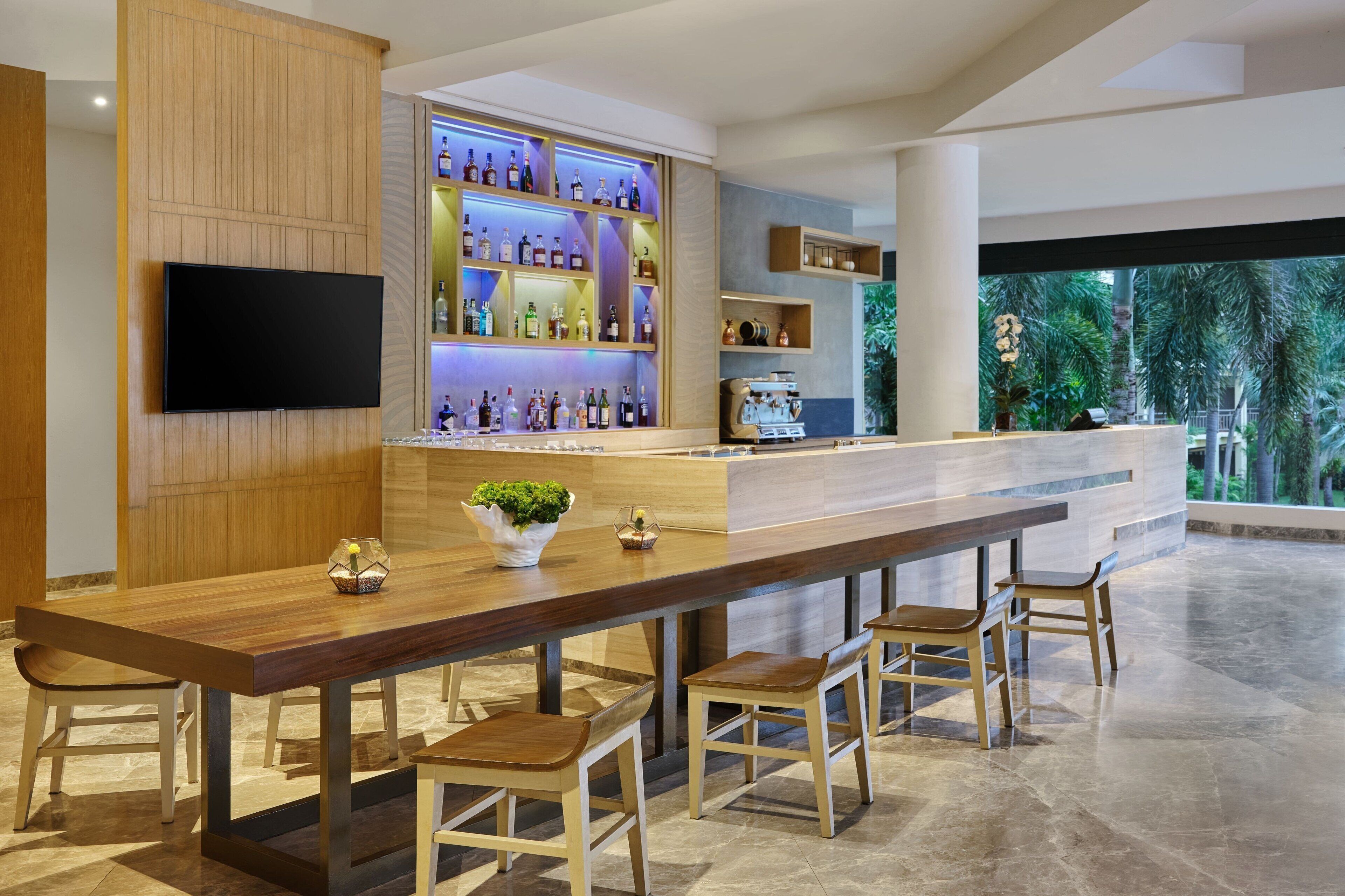 A bar area with a long wooden counter, multiple stools, a television mounted on the wall, and shelves with bottles.