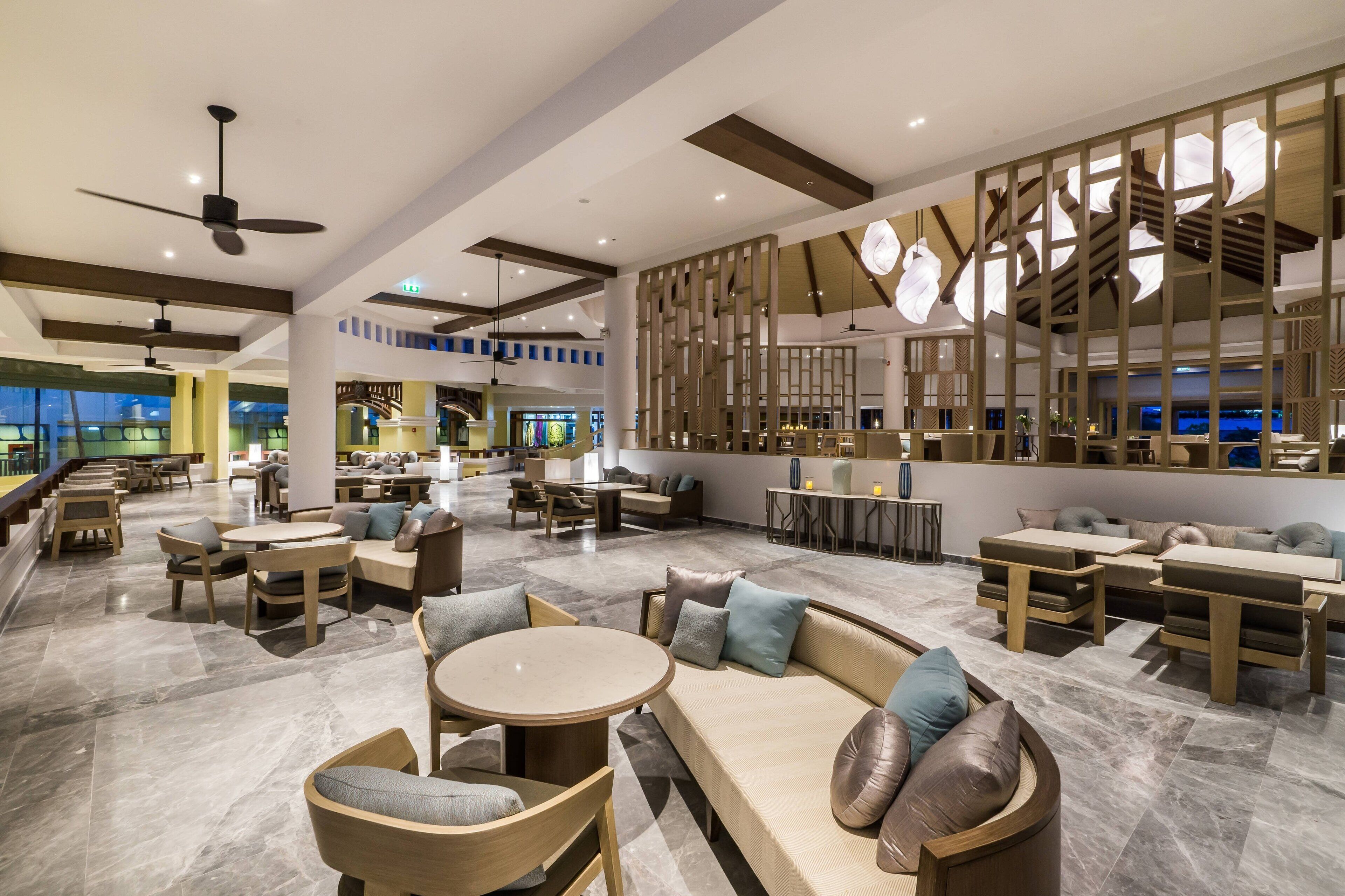 Un hall d’hôtel spacieux, doté de plusieurs espaces de détente, notamment des canapés et des tables, ainsi qu’un grand mur décoratif orné de motifs géométriques.