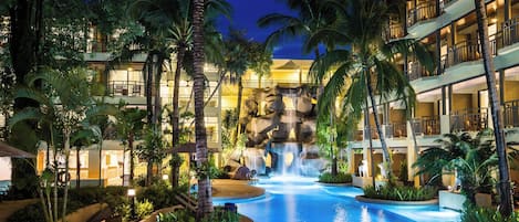 A resort pool with a waterfall, surrounded by palm trees and a multi-story building.