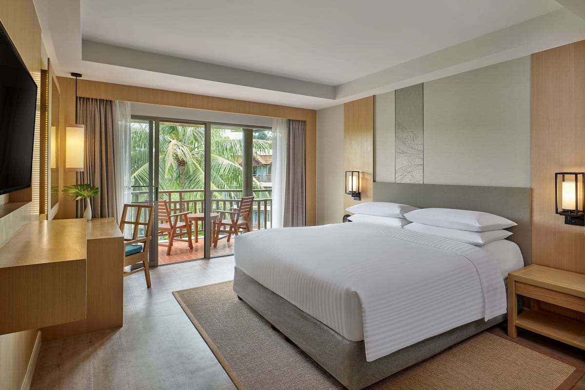 A hotel room with a large bed, a desk, a TV, and a balcony with a view of palm trees.