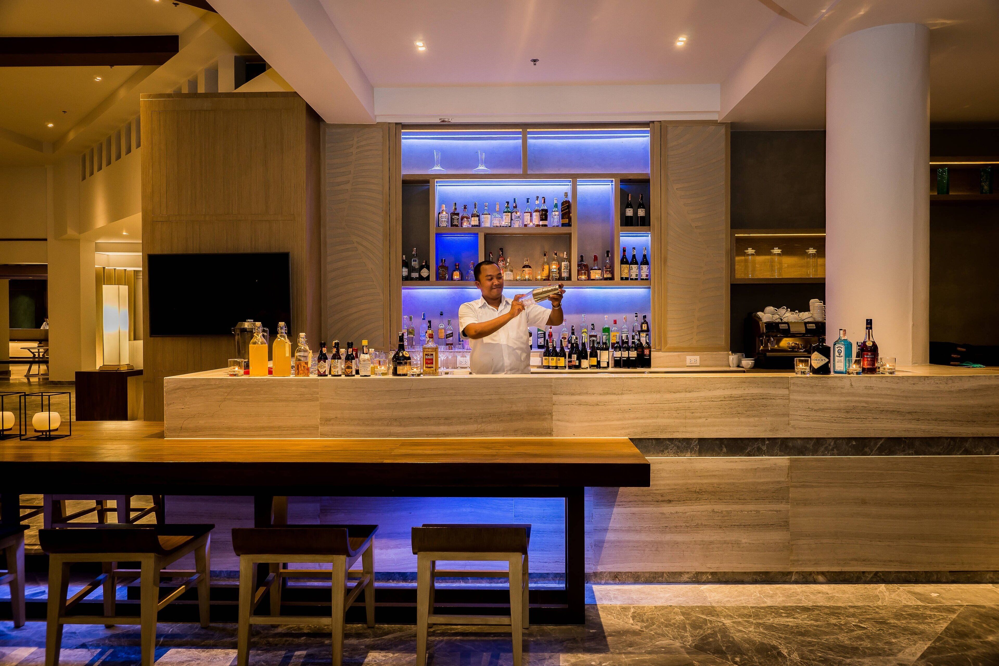 Zona de bar moderna con un bartender, una estantería llena de licores y asientos.