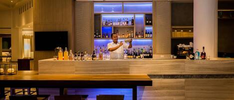 Zona de bar moderna con un bartender, una estantería llena de licores y asientos.