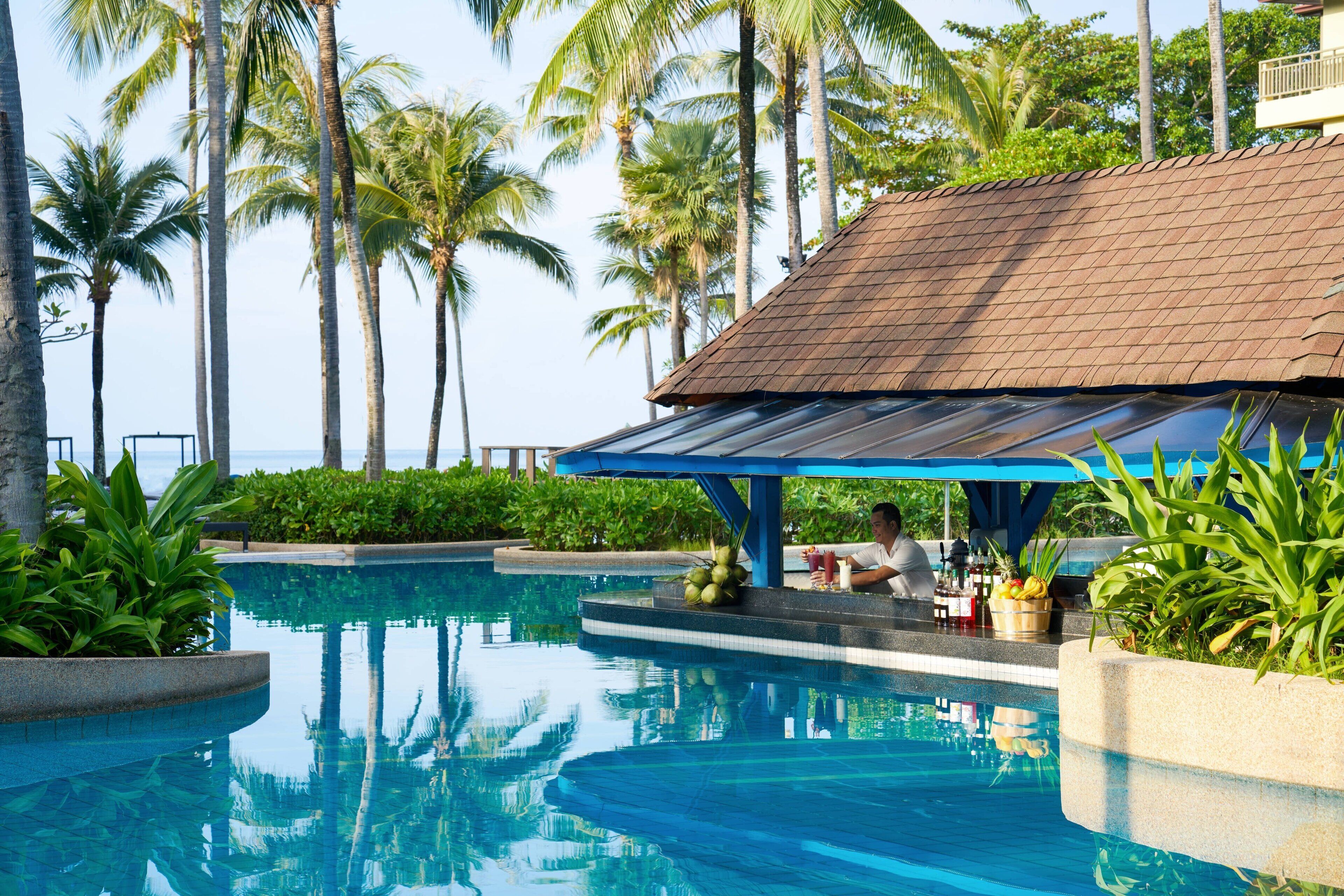 A pool area with a person relaxing under a shaded structure, surrounded by palm trees and a clear blue sky.