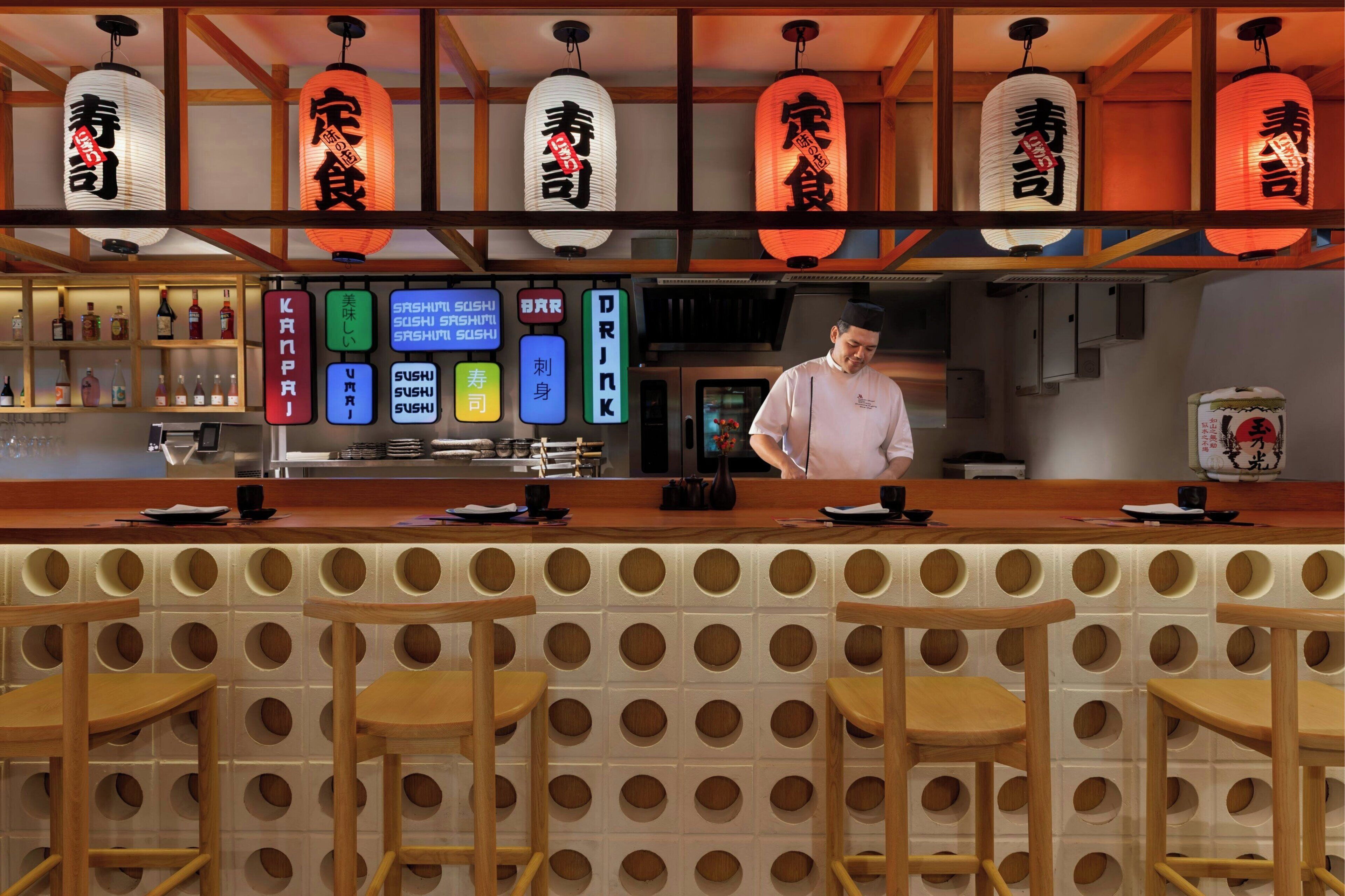 A sushi bar with a chef, wooden stools, and illuminated signs indicating menu items.