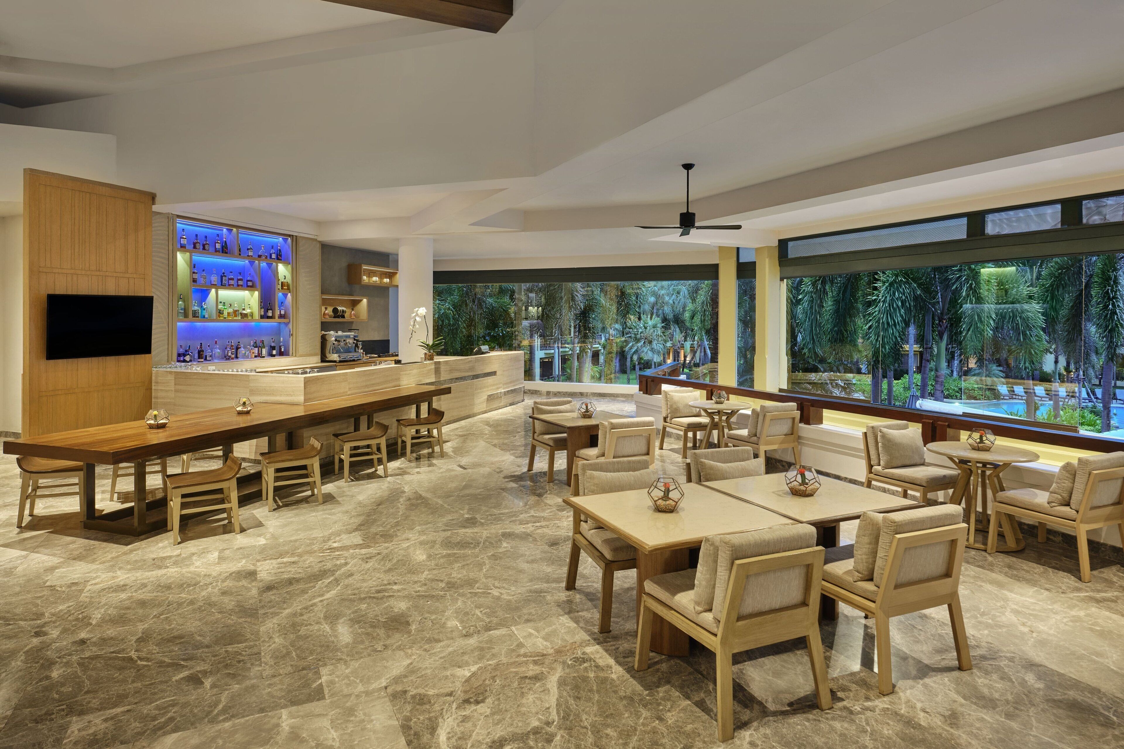 A modern bar area with a long counter, seating with cushioned chairs, and a large window offering a view of palm trees and a pool.