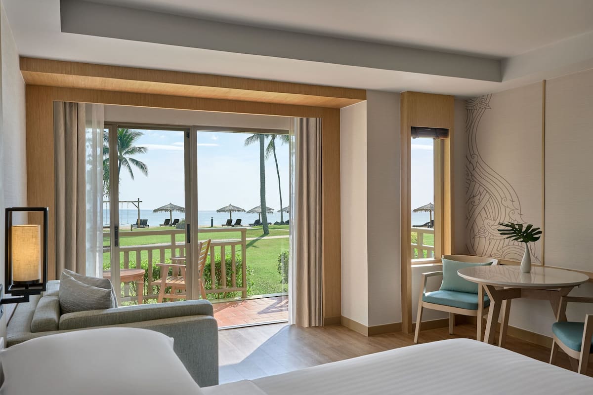 A hotel room with a large sliding glass door leading to a balcony with a view of palm trees and the sea. There is a bed, a small table, and a chair on the balcony.