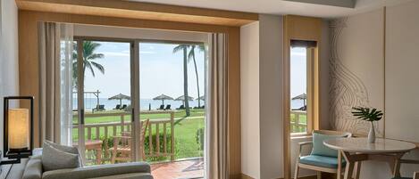 A hotel room with a large sliding glass door leading to a balcony with a view of palm trees and the sea. There is a bed, a small table, and a chair on the balcony.