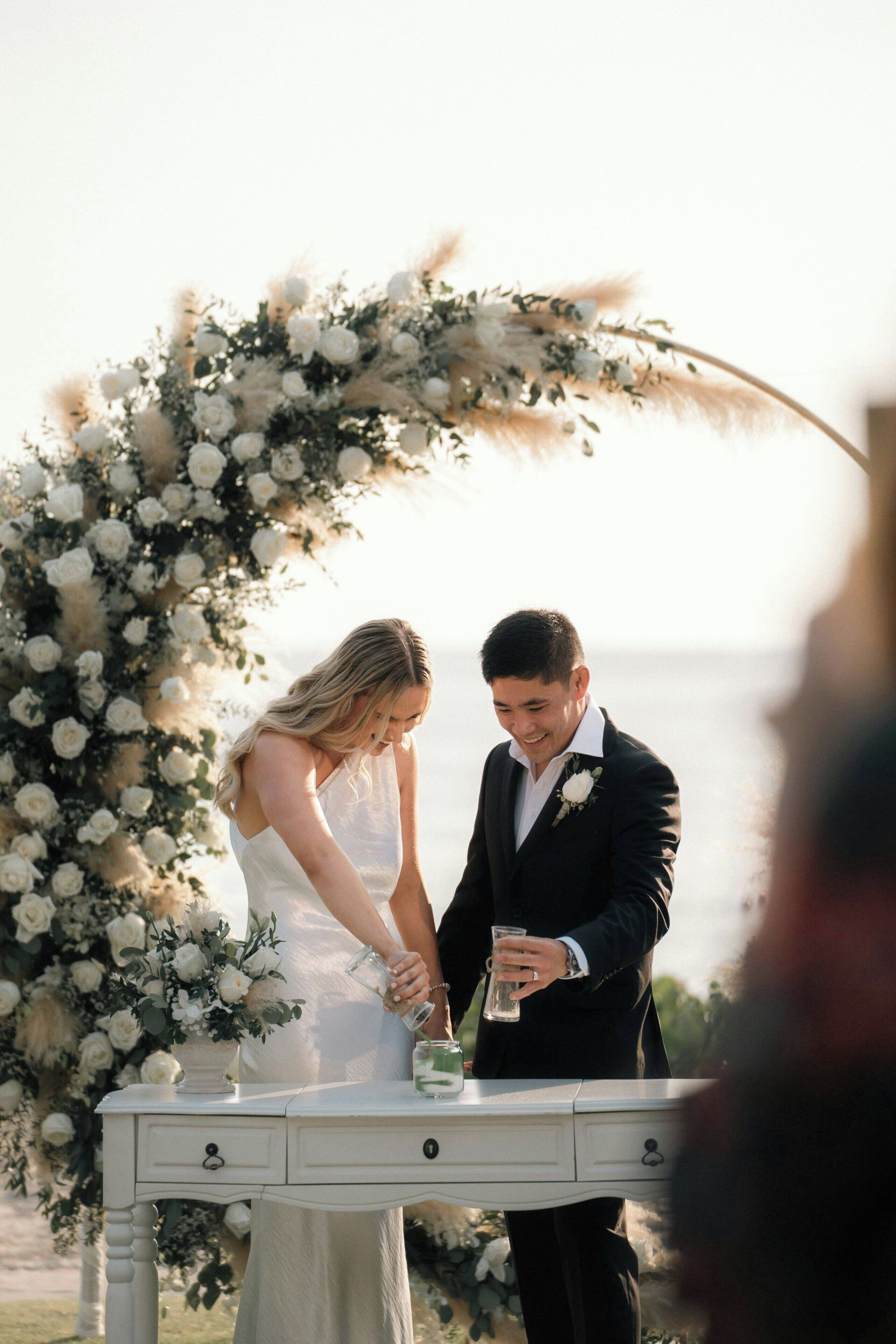 A wedding ceremony with a white floral arch and a table with a glass of water.