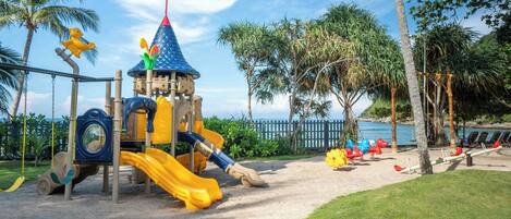 A playground with slides and climbing structures, surrounded by palm trees and a view of the ocean.