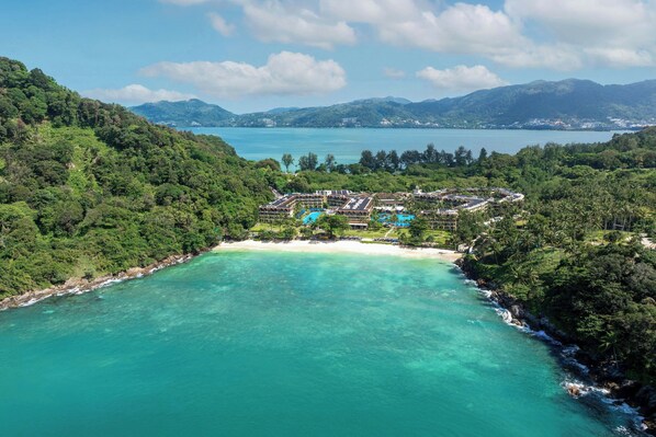An aerial view of a resort with a large pool, surrounded by lush greenery and a beach.
