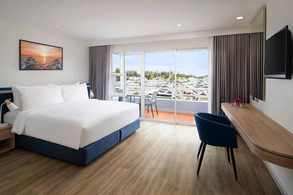 Quarto de hotel com uma cama grande, uma escrivaninha, uma cadeira, uma televisão e uma varanda com vista para barcos.
