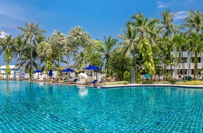 Una piscina con sillones de descanso y sombrillas, rodeada de palmeras y un edificio de fachada blanca.