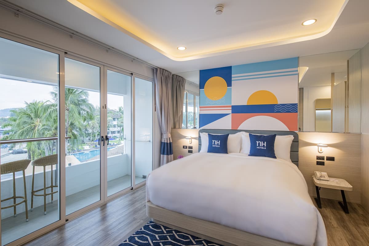 Quarto de hotel moderno com uma cama grande, varanda com cadeiras de vime, vista para palmeiras e um mural de parede abstrato e colorido.