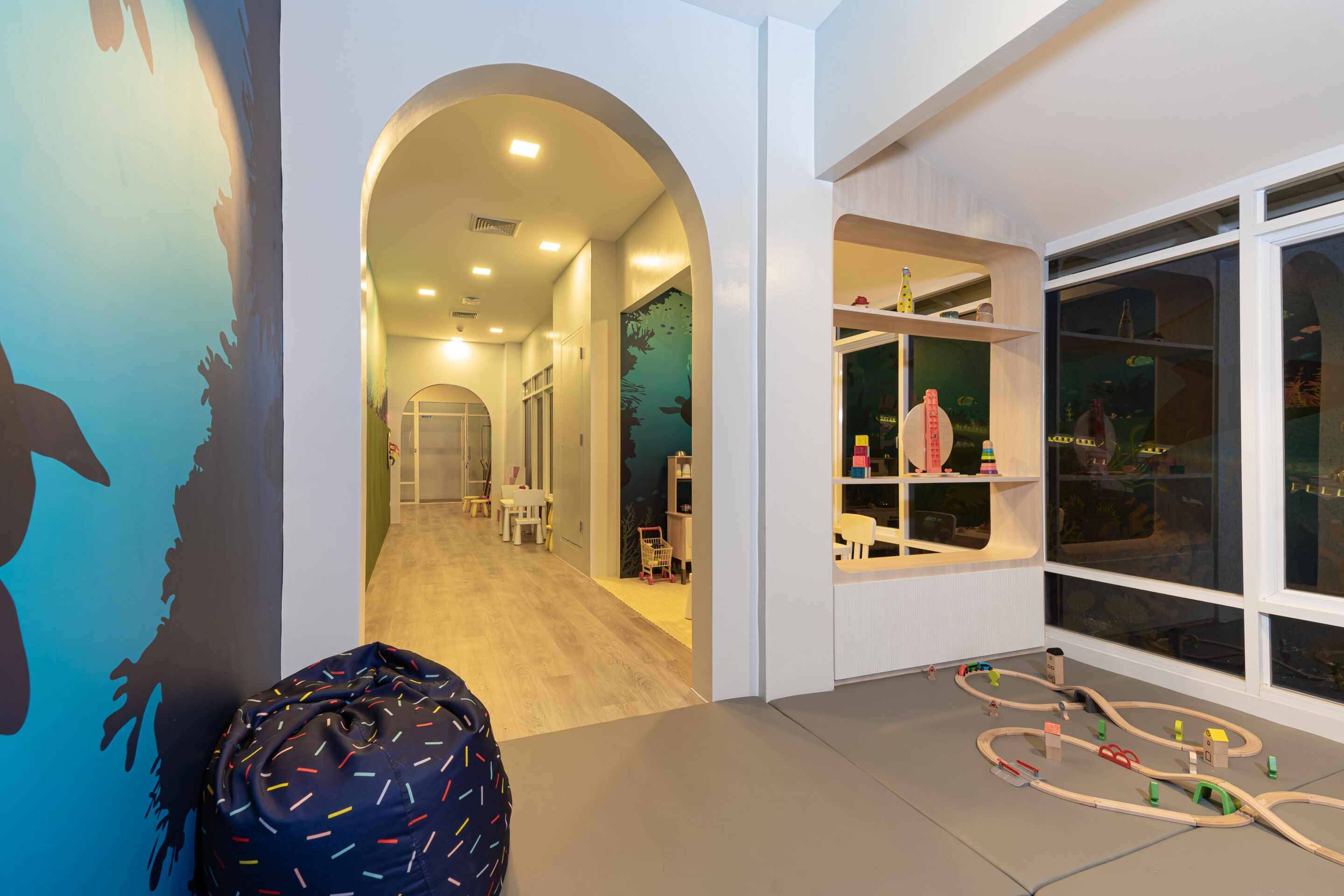 A children's play area with a train track layout, a bean bag, and a mural on the wall.
