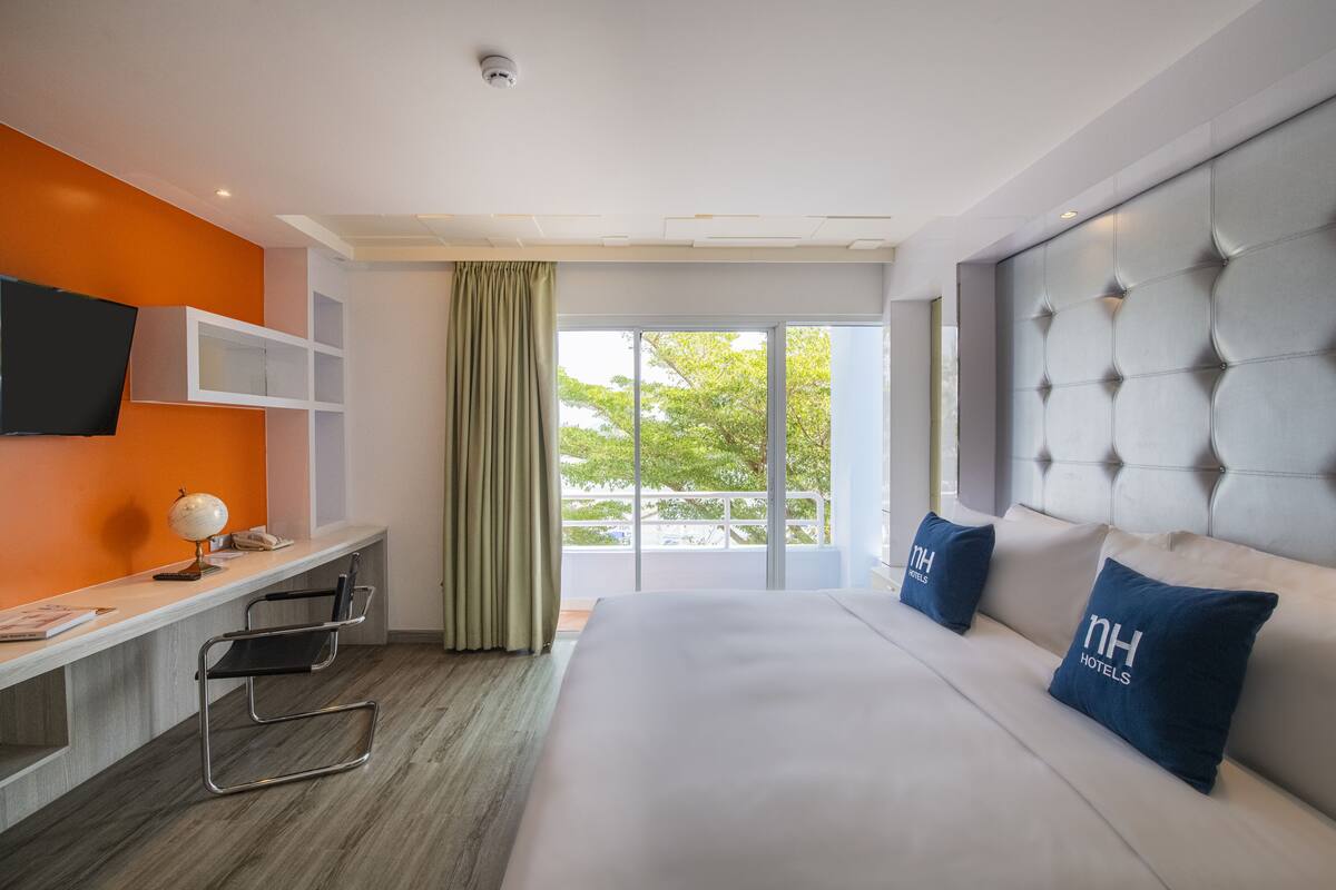 Quarto de hotel moderno com uma cama grande, uma escrivaninha com um globo, uma televisão e varanda com vista para árvores.