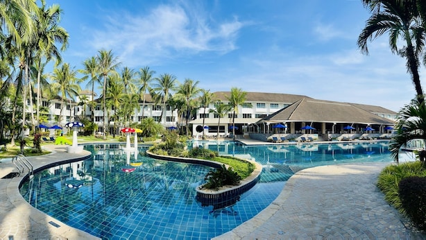 A large outdoor swimming pool with a curved design, surrounded by palm trees and lounge areas with umbrellas. - NH Boat Lagoon Phuket Resort (Ko Kaeo)