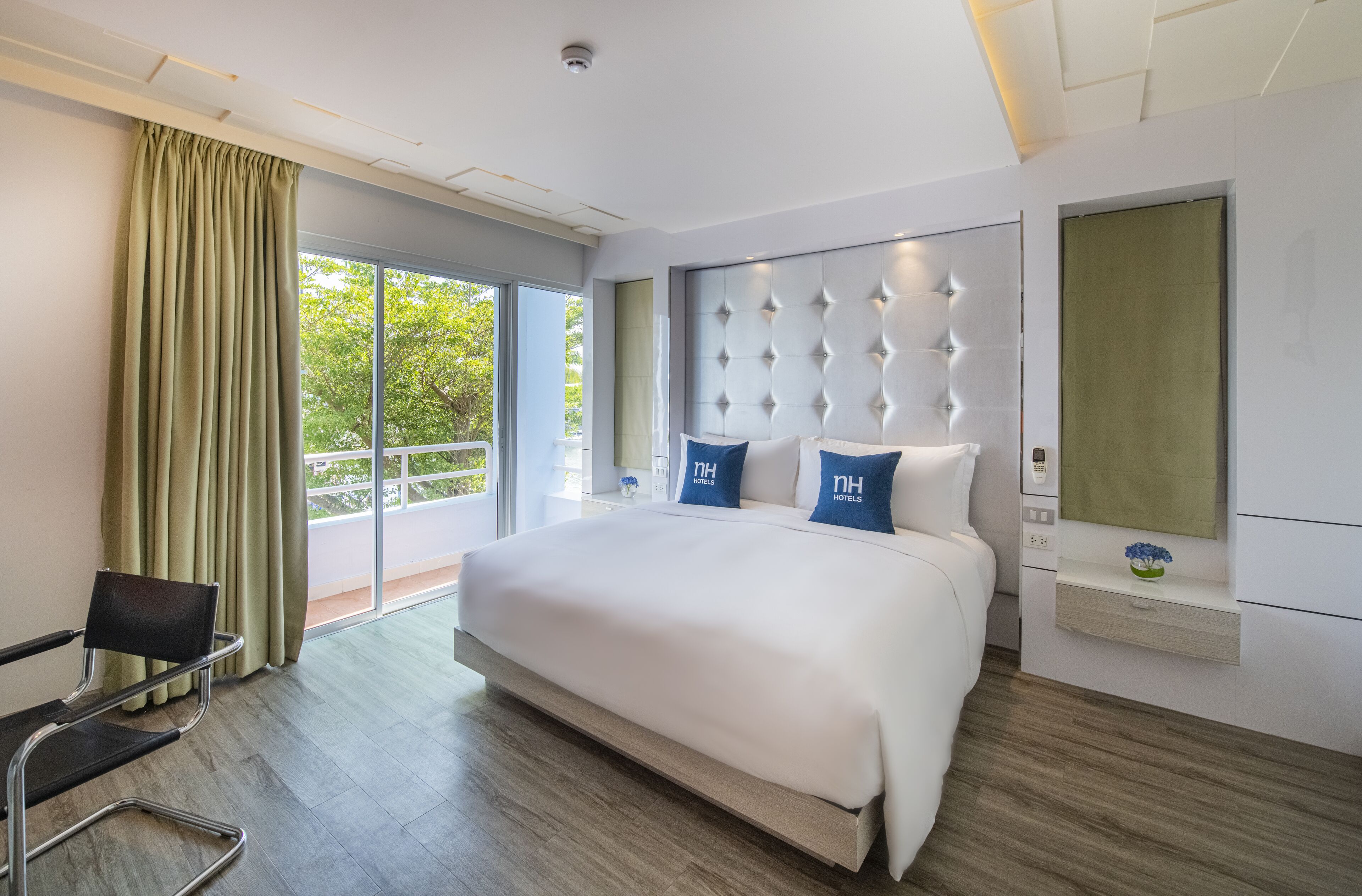 A modern hotel room with a large bed, a chair, a window with curtains, and a view of greenery outside.