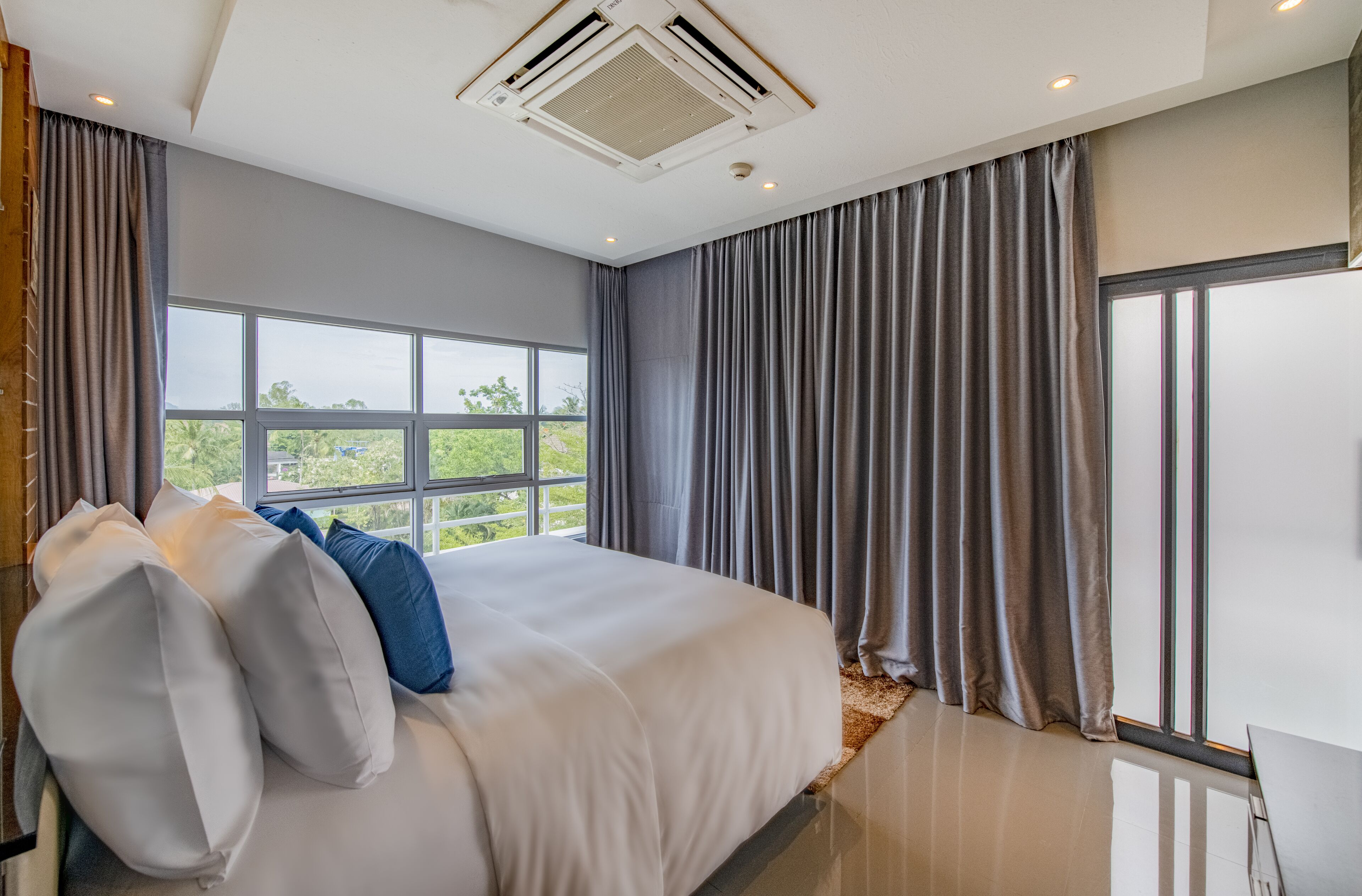A modern bedroom with a large bed, grey curtains, and a view of greenery outside.
