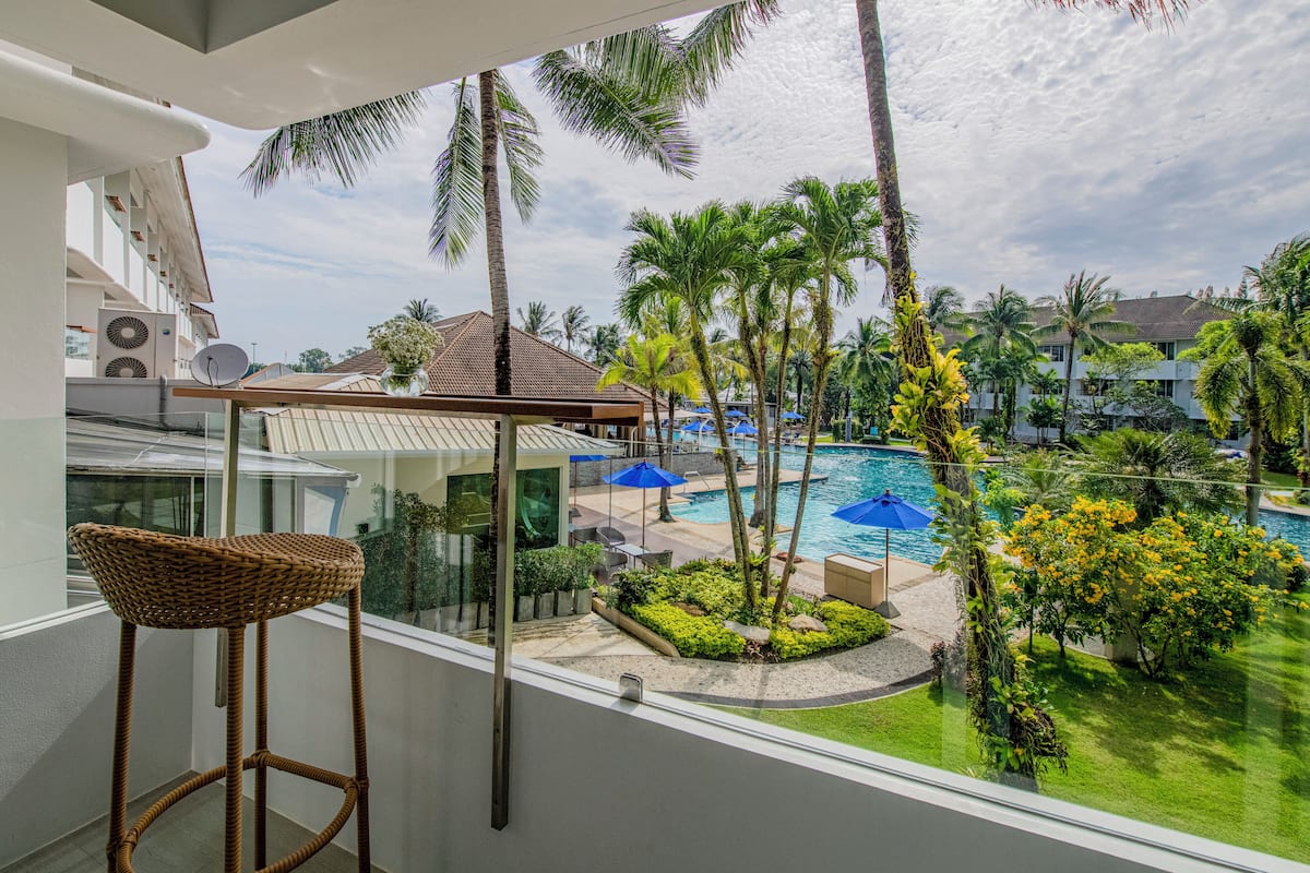 Uma varanda com cadeira de vime, com vista para uma área de piscina com palmeiras e céu claro.