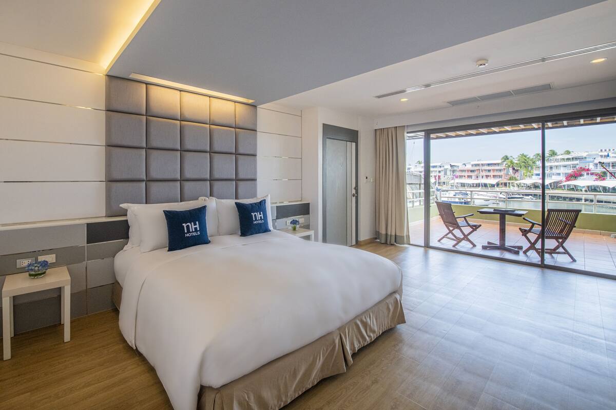 Quarto de hotel moderno com uma cama grande, uma mesinha de cabeceira, vista da cidade através de amplas janelas e uma varanda com área de estar externa.