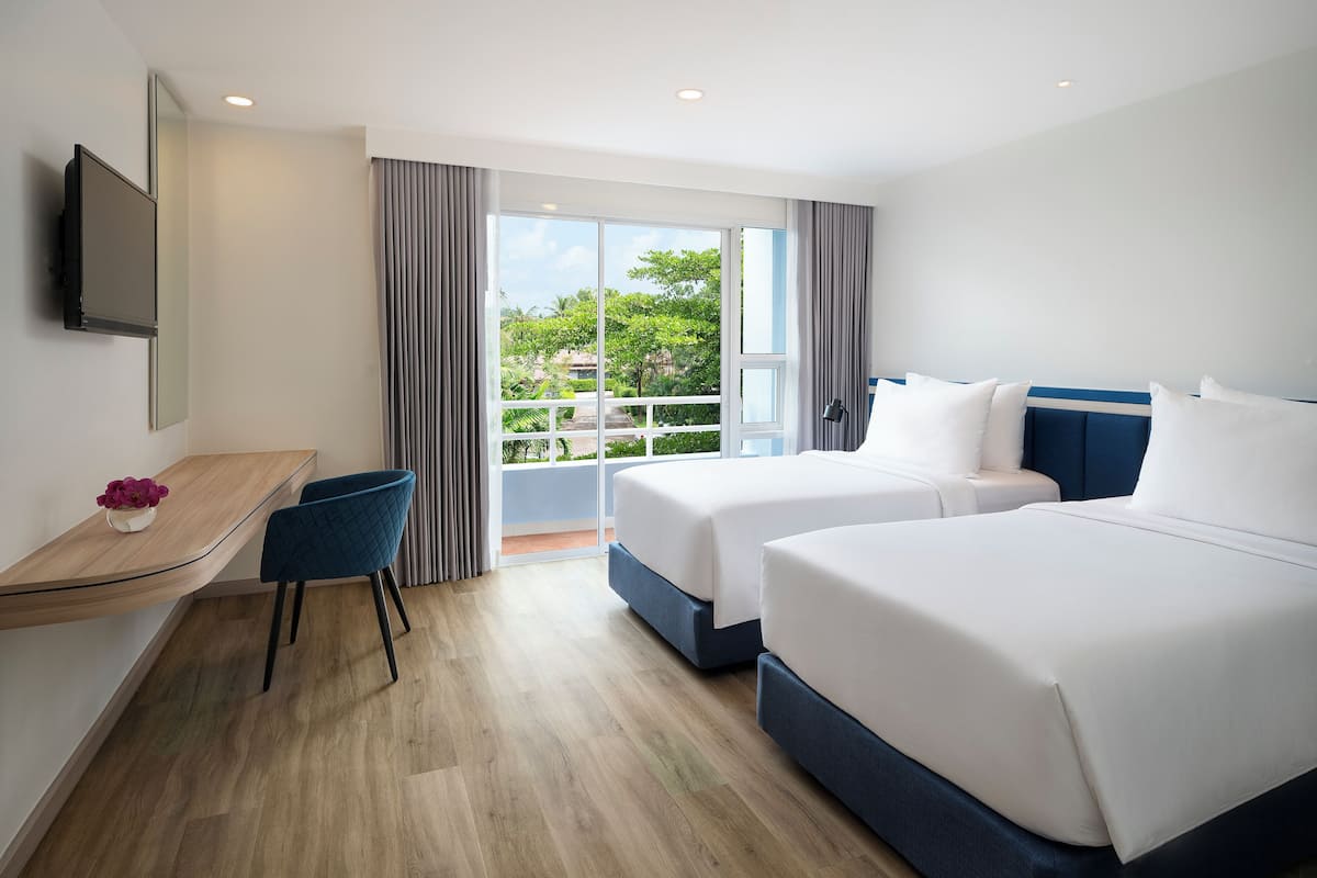 Quarto de hotel com duas camas, uma escrivaninha, uma cadeira, televisão e varanda com vista para o verde.