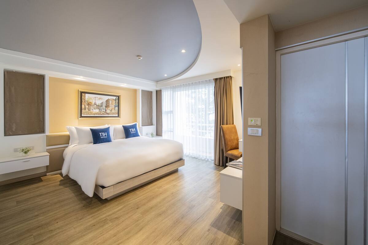 Quarto de hotel moderno com uma cama grande, uma mesa de cabeceira, uma cadeira, um quadro na parede e uma janela com cortinas.