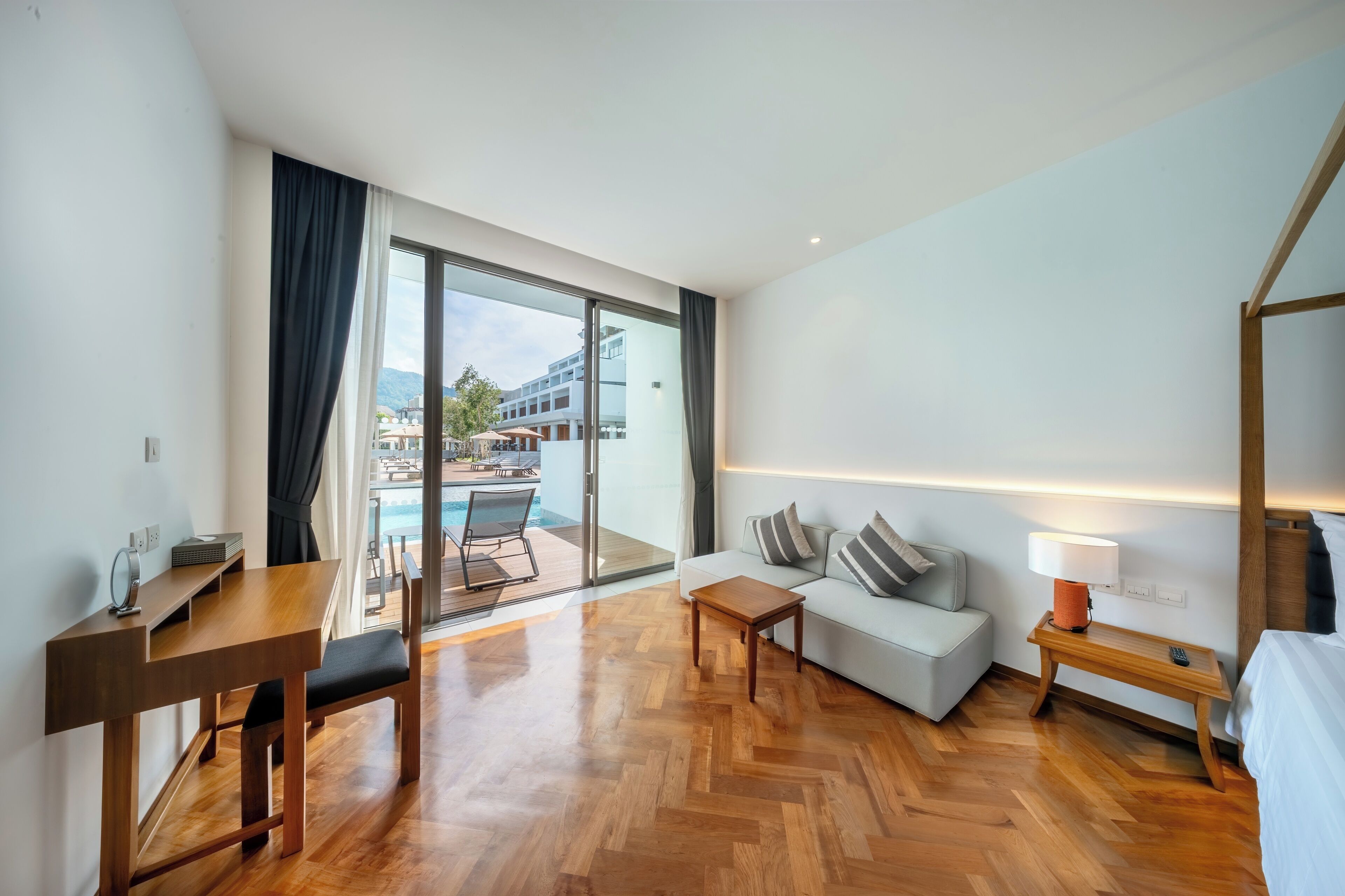 A modern hotel room with a wooden floor, a sofa, a table, a lamp, and a view of a swimming pool and buildings.