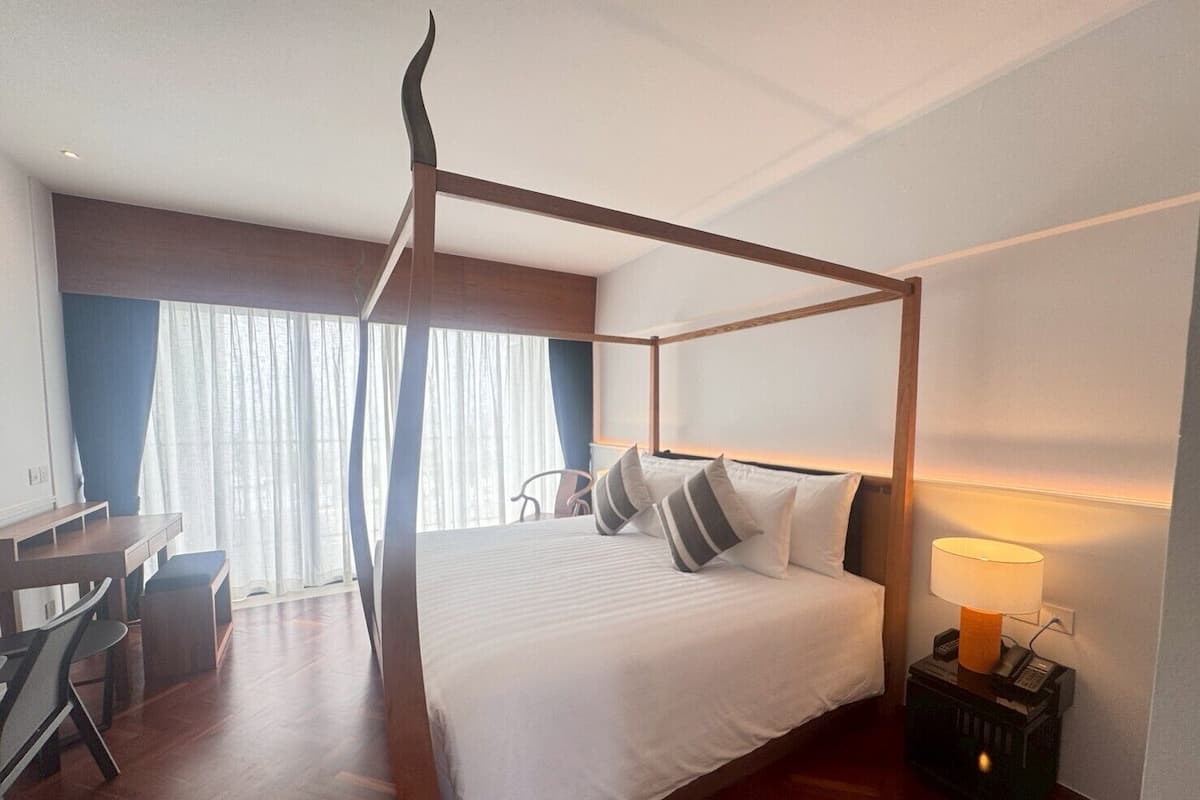 A modern hotel room with a four-poster bed, a desk with a chair, and a bedside table with a lamp.
