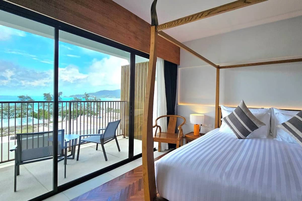 A bedroom with a large bed, a balcony with a table and chairs, and a view of the ocean.