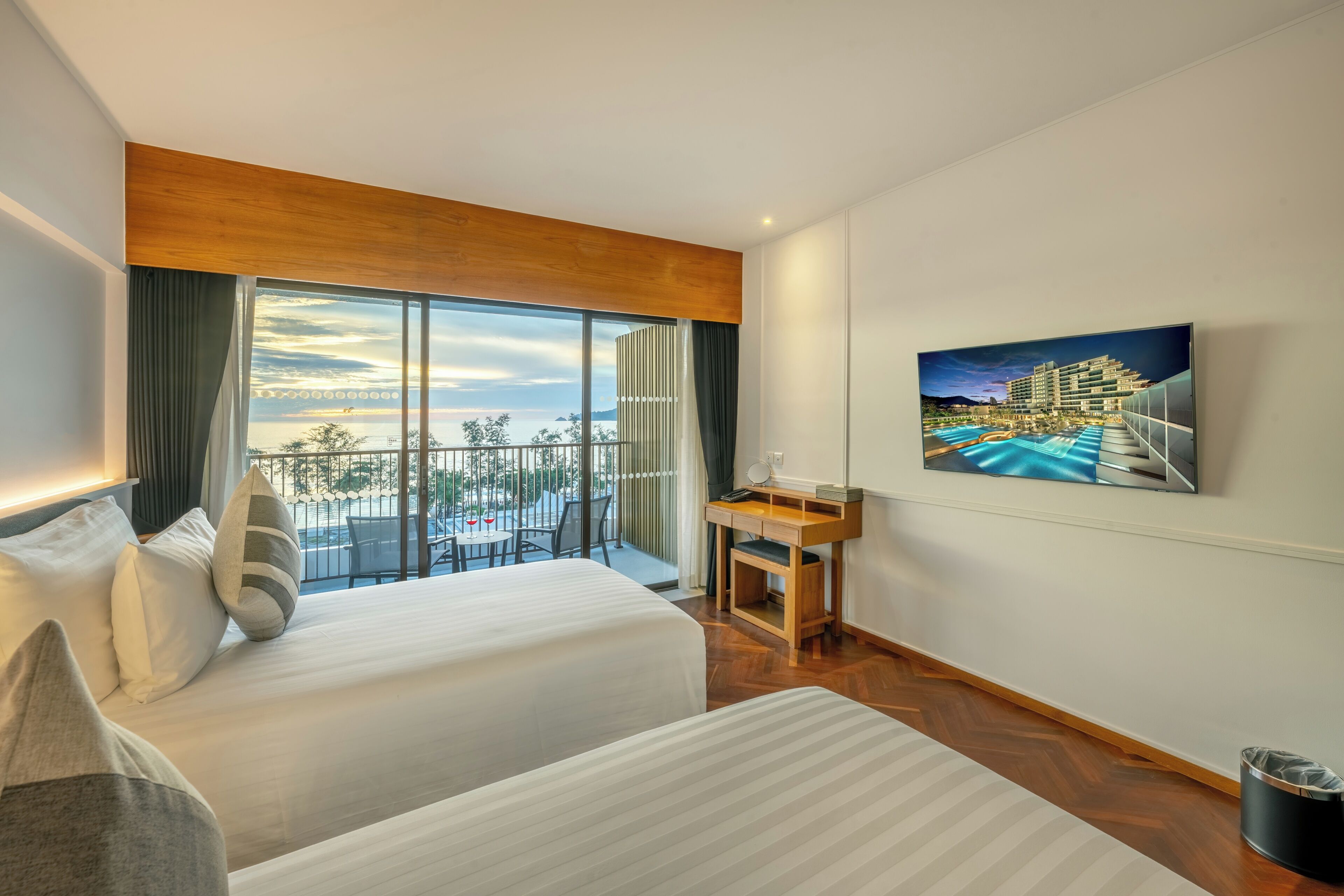 A hotel room with two beds, a desk, a large window with a view of a pool and building, and a painting on the wall.