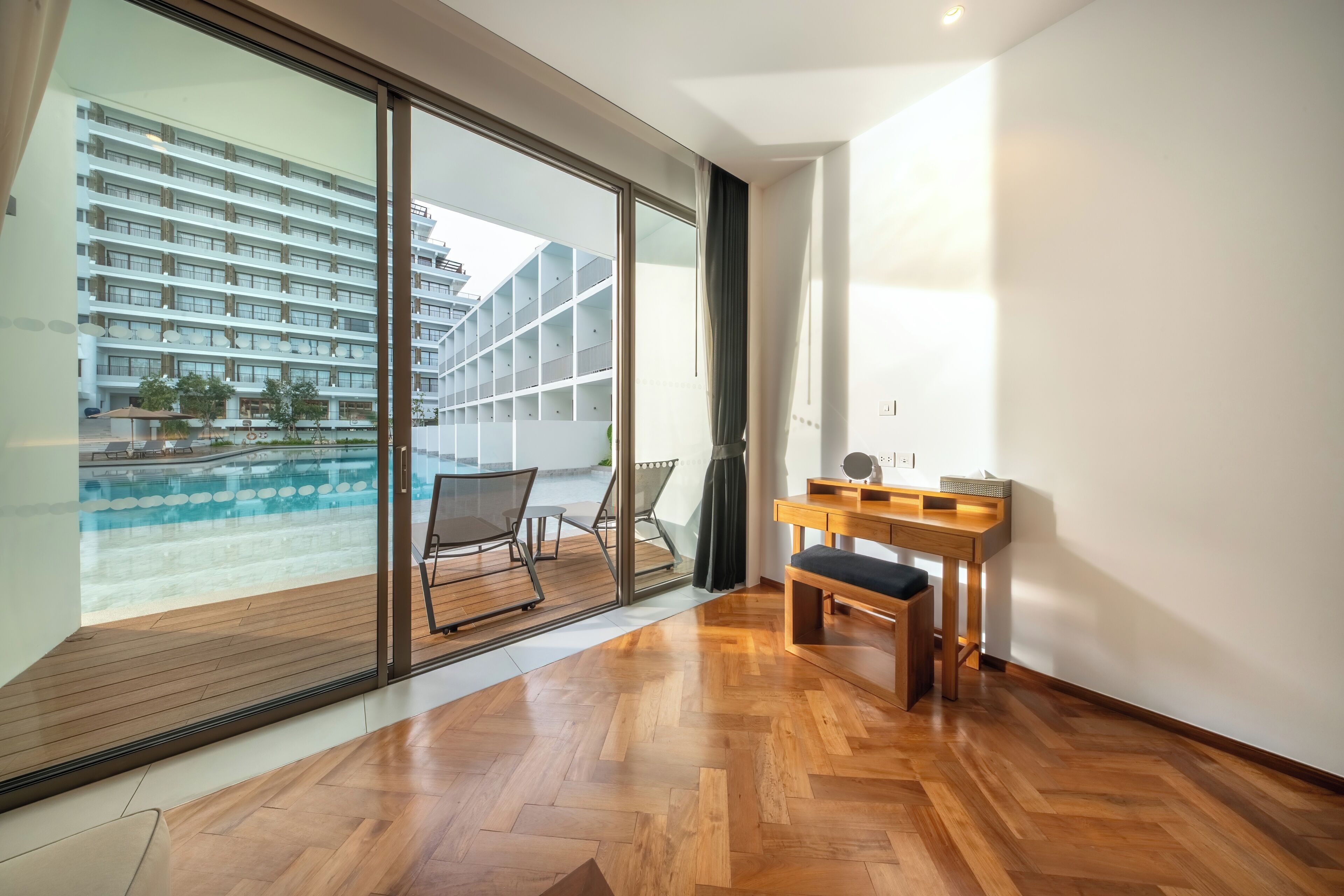 Una camera moderna con pavimento in legno, un'ampia finestra vista piscina e palazzi, una scrivania in legno e una sedia.