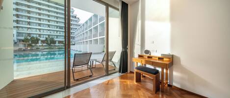 Una camera moderna con pavimento in legno, un'ampia finestra vista piscina e palazzi, una scrivania in legno e una sedia.