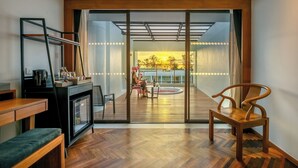 A modern living room with a wooden herringbone floor, a black entertainment unit, a wooden bench, and a glass door leading to a balcony with a view of a sunset. - Patong Beach Hotel by See2Sea (Patong)