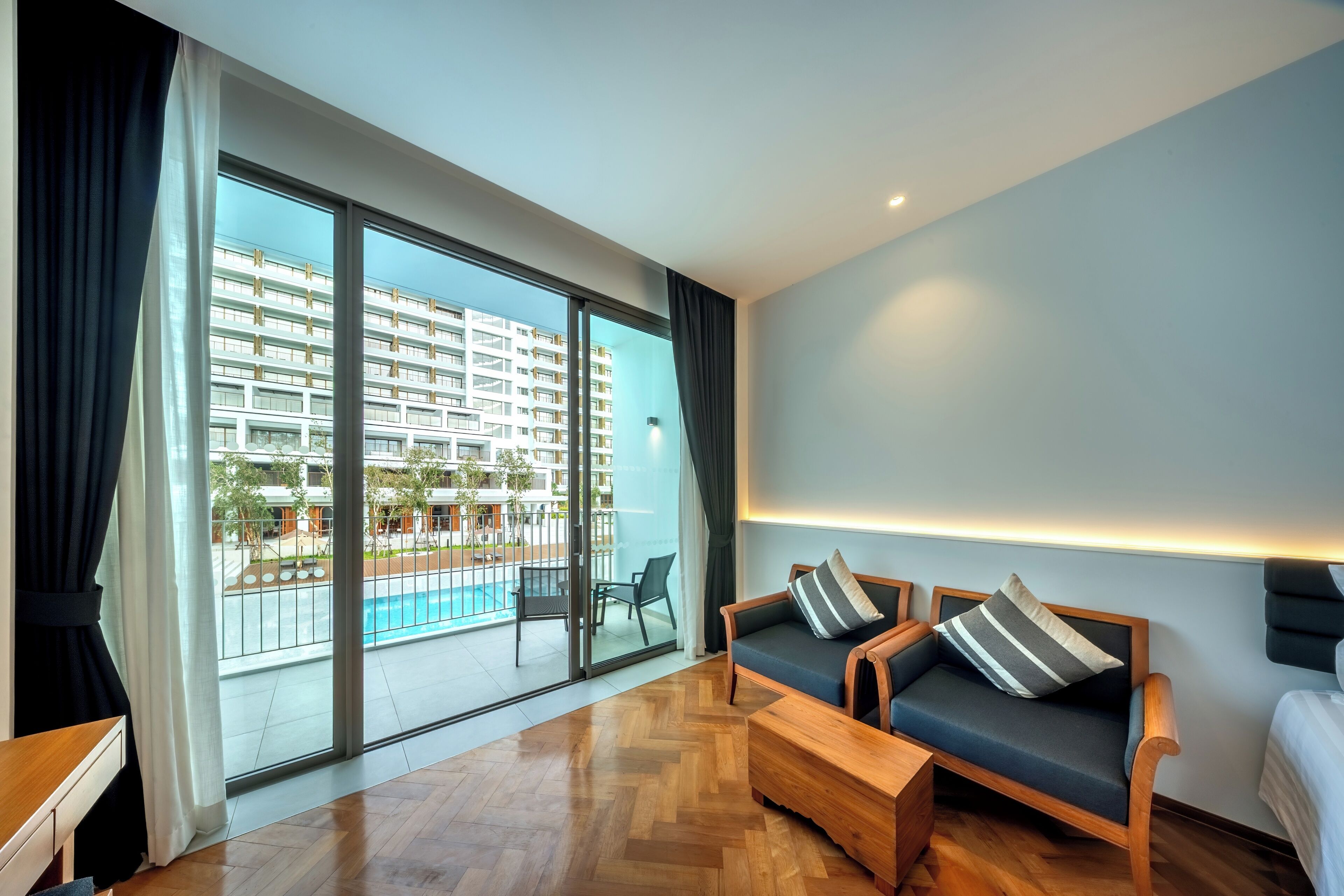 A modern hotel room with a large sliding glass door, a wooden floor, a sofa with striped pillows, and a bed with a dark headboard.