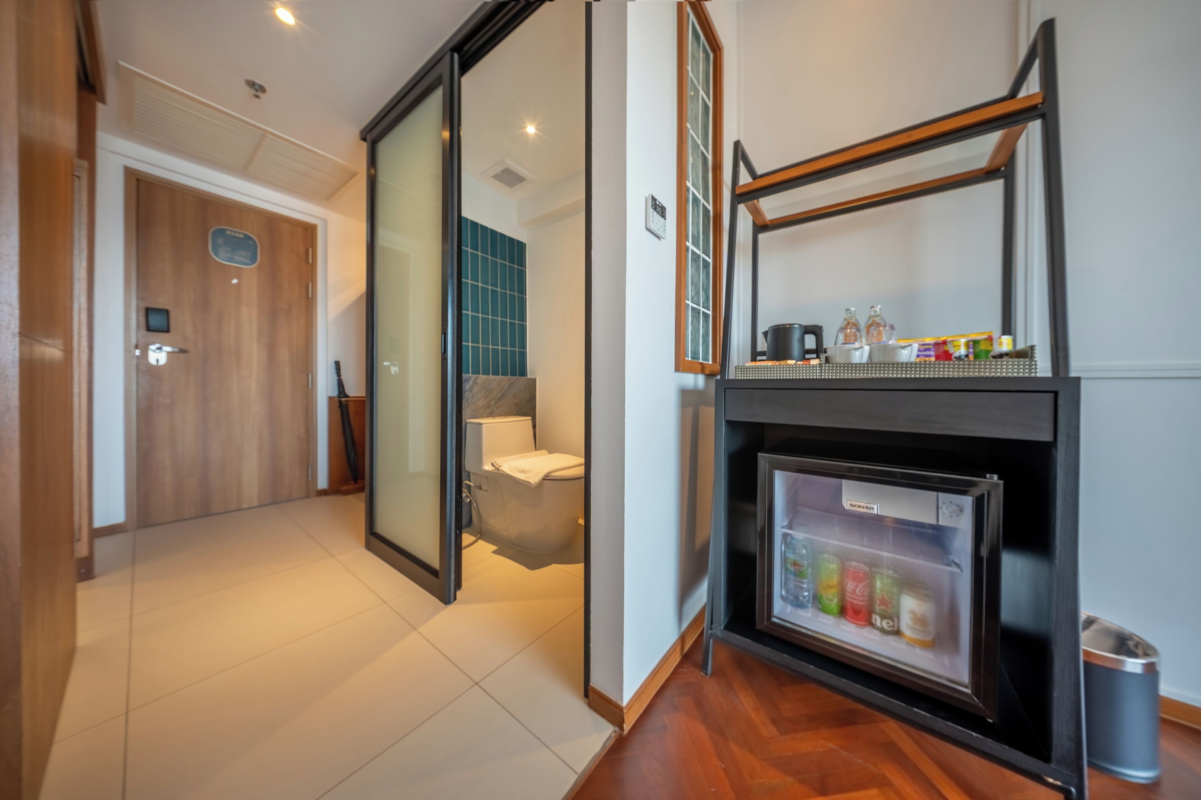 A hotel room with a sliding door leading to a bathroom, a small refrigerator with beverages, and a shelf with bottles.