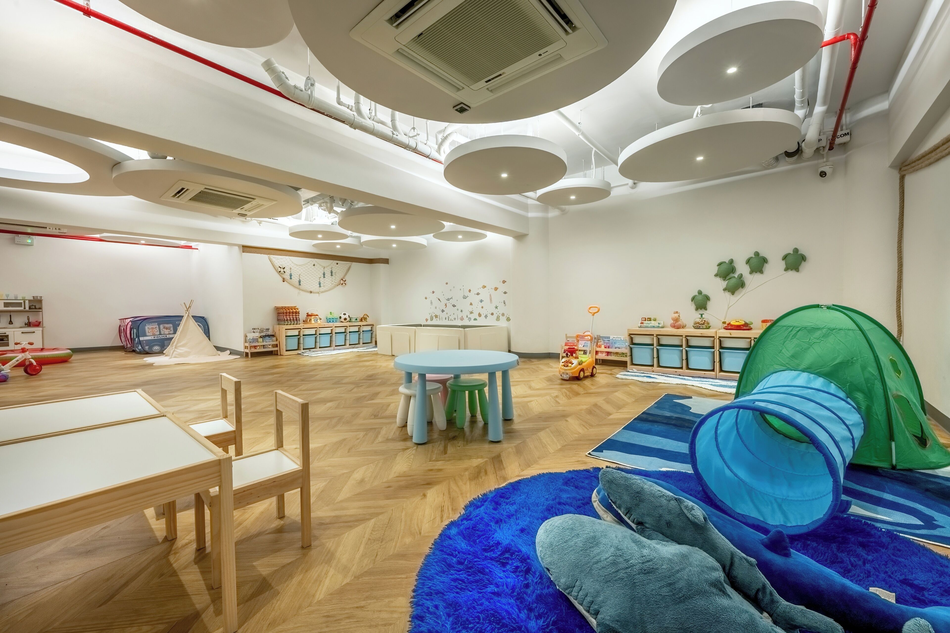 Um quarto espaçoso com teto alto, luminárias circulares no teto e um espaço de brincadeira com uma tenda, além de um tapete azul.
