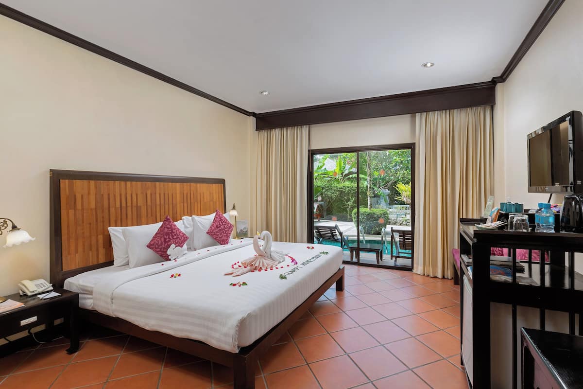 A hotel room with a large bed, a desk with a TV, and a balcony with a view of greenery.