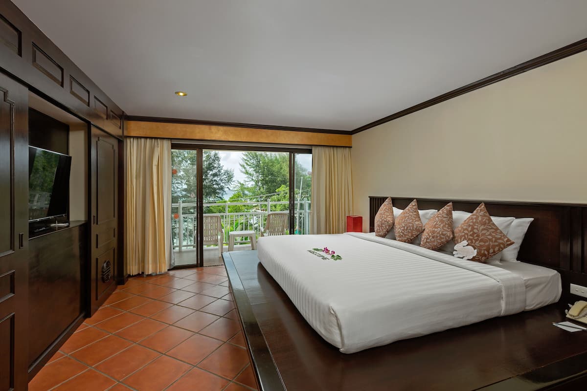 A hotel room with a large bed, a TV, and a balcony with a view of trees.