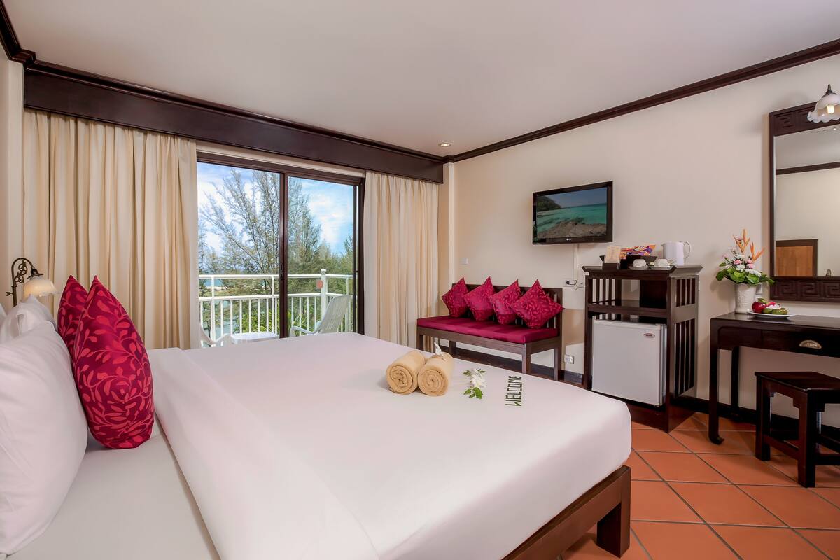 A hotel room with a bed, a desk, a chair, a TV, and a balcony with a railing.