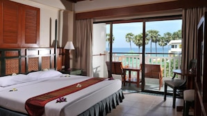 A hotel room with a large bed, a desk, a chair, and a balcony with a view of the ocean and palm trees. - Karon Princess Hotel (Karon)