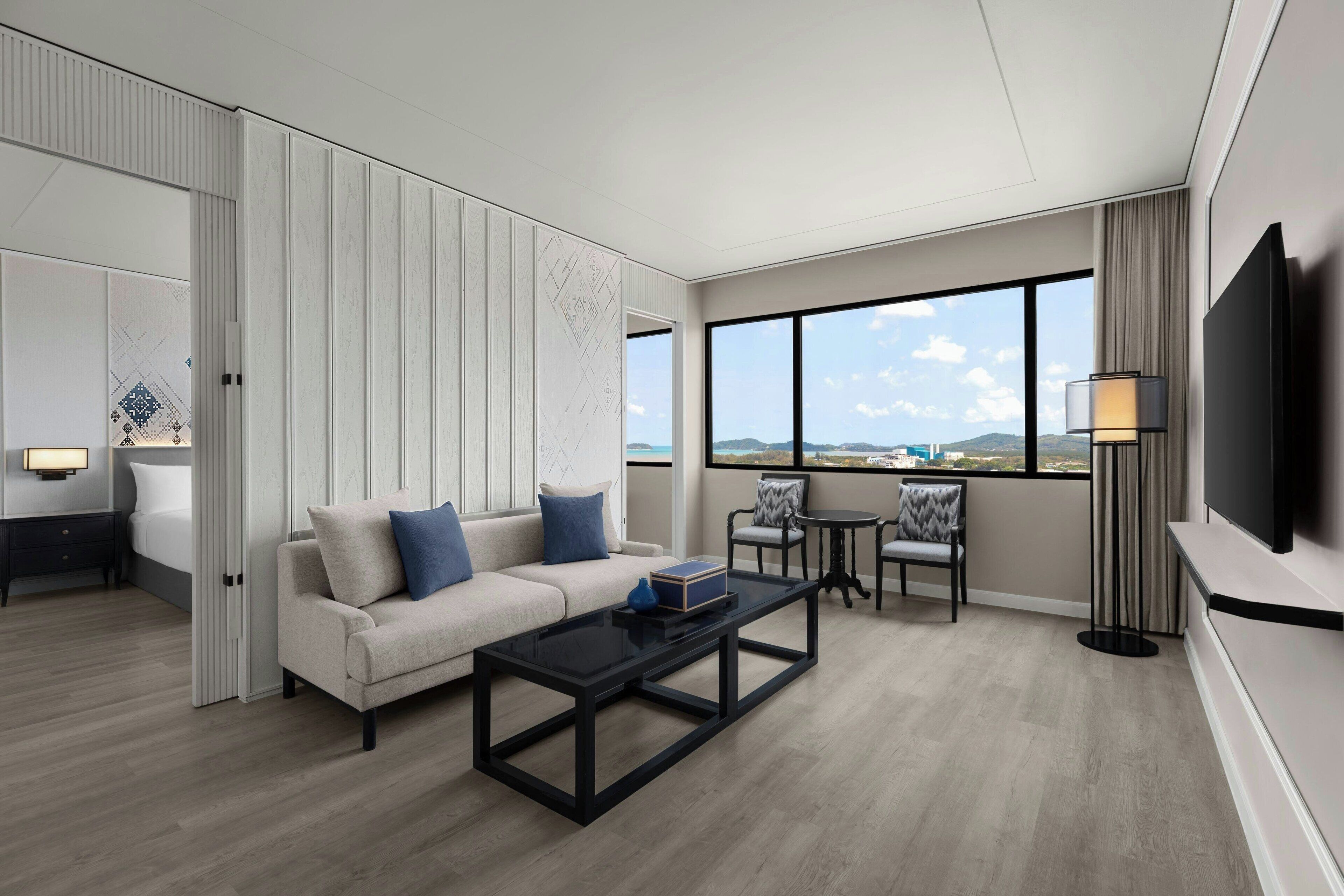 Habitación de hotel moderna con sofá, mesa de centro y sillas. Tiene un televisor montado en la pared y un ventanal con vistas a la ciudad.