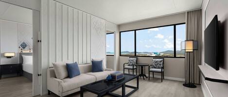 A modern hotel room with a sofa, coffee table, and chairs. There is a TV mounted on the wall and a large window offering a view of the city.