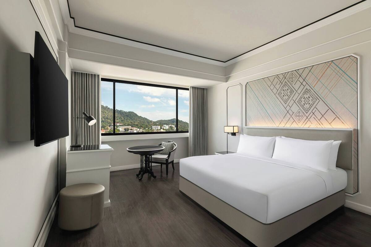 Une chambre d’hôtel moderne dotée d’un grand lit, d’un téléviseur à écran plat, d’une petite table avec une chaise et offrant une vue sur la ville grâce à de grandes fenêtres.