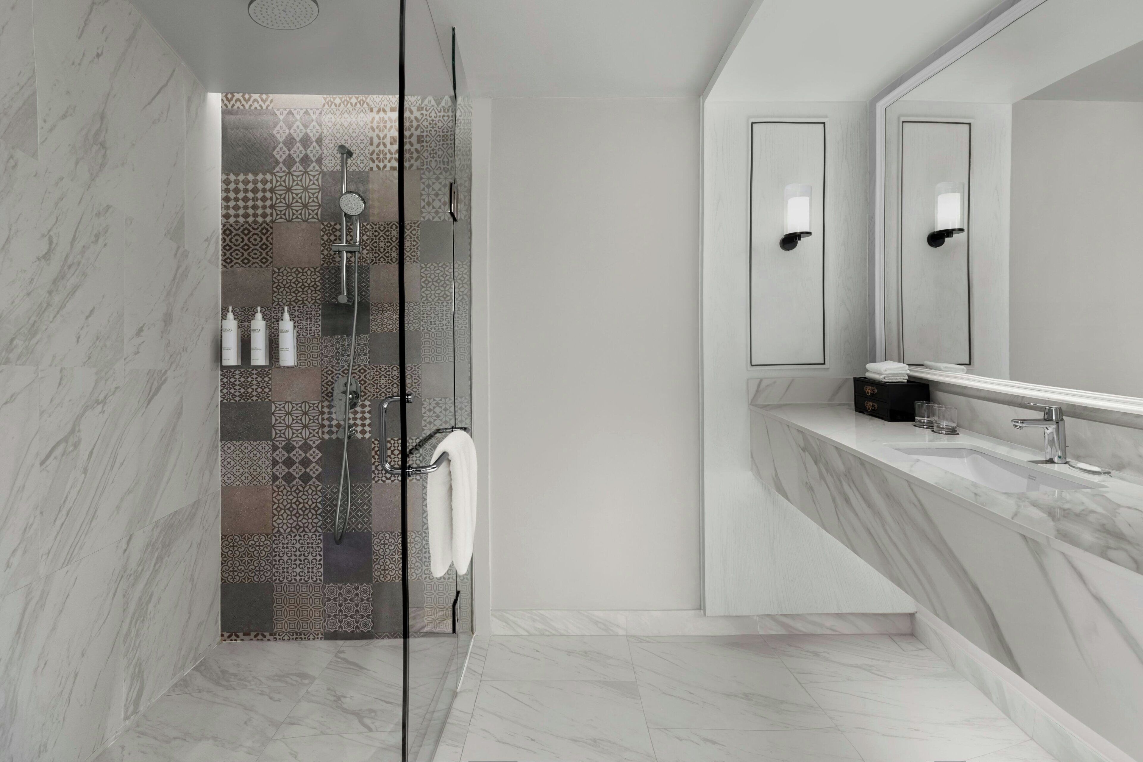 Een moderne badkamer met een douchegedeelte voorzien van patroon betegelde tegels, een douche met glazen wand, een wastafel met marmeren blad en een grote spiegel.